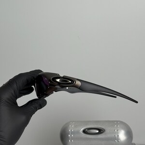 May include: Black and gray sports sunglasses with a sleek, modern design. The sunglasses have a dark lens and a curved frame. The Oakley logo is visible on the frame. The sunglasses are held by a gloved hand.