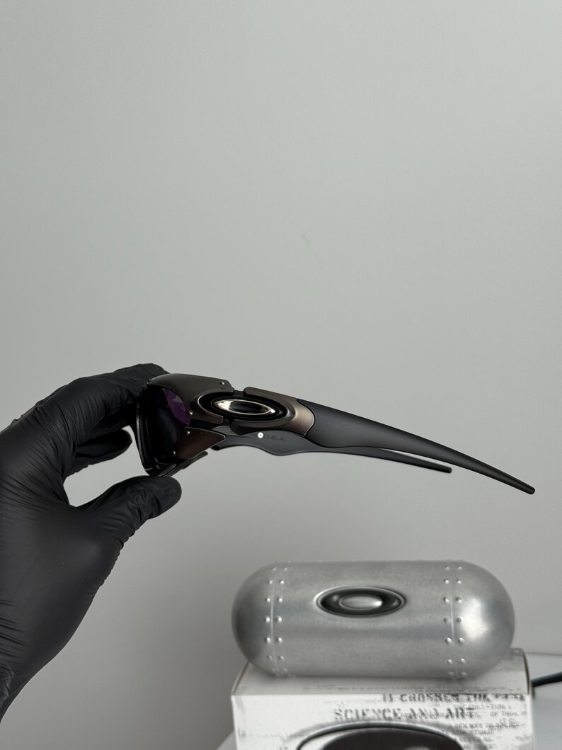 May include: Black and gray sports sunglasses with a sleek, modern design. The sunglasses have a dark lens and a curved frame. The Oakley logo is visible on the frame. The sunglasses are held by a gloved hand.