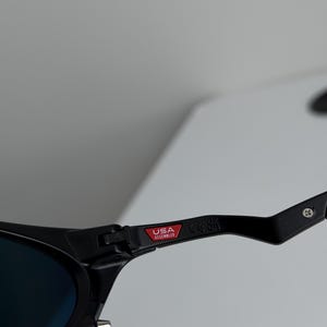 May include: Close-up of black sunglasses with blue lenses. The frame features a red tag that reads "USA Assembled". The sunglasses have a sleek, modern design.