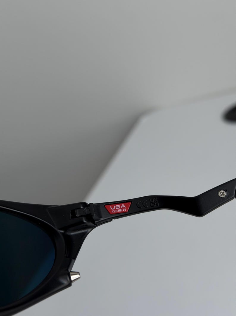 May include: Close-up of black sunglasses with blue lenses. The frame features a red tag that reads "USA Assembled". The sunglasses have a sleek, modern design.