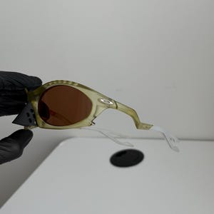 May include: Olive green sunglasses with brown lenses. The frames have a textured pattern and the arms are white. The sunglasses are held by a gloved hand.