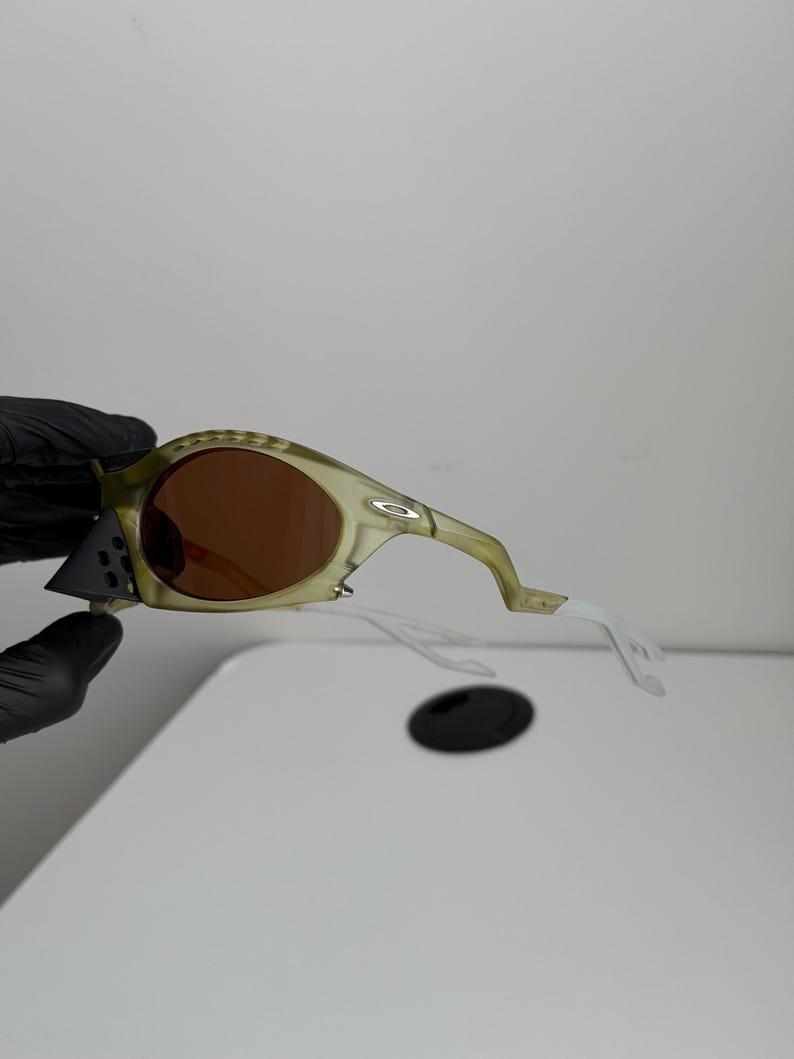 May include: Olive green sunglasses with brown lenses. The frames have a textured pattern and the arms are white. The sunglasses are held by a gloved hand.