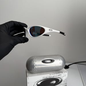 May include: White sunglasses with a curved frame and blue-tinted lenses. The sunglasses are held above a silver case and a white box with the text "SCIENCE AND ART". The sunglasses have black accents on the arms.