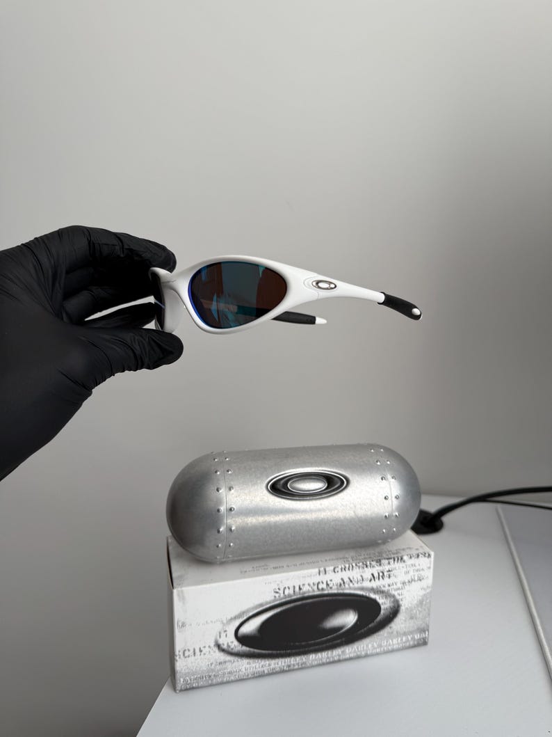 May include: White sunglasses with a curved frame and blue-tinted lenses. The sunglasses are held above a silver case and a white box with the text "SCIENCE AND ART". The sunglasses have black accents on the arms.