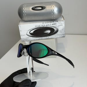 May include: Black sunglasses with green lenses are displayed on a clear stand. The sunglasses have a sleek, modern design. A silver case and a box with the text "SCIENCE AND ART" are in the background.