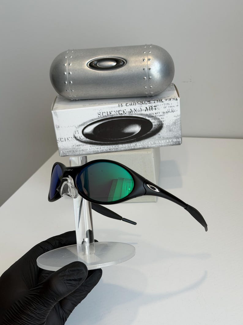 May include: Black sunglasses with green lenses are displayed on a clear stand. The sunglasses have a sleek, modern design. A silver case and a box with the text "SCIENCE AND ART" are in the background.