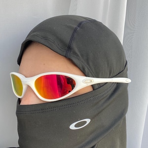 Puede incluir: Gafas de sol con montura blanca y lentes espejadas rojas. Las gafas se llevan con un pasamonta&ntilde;as negro con el logotipo blanco de Oakley. Las lentes reflejan un vibrante tono rojo y amarillo.