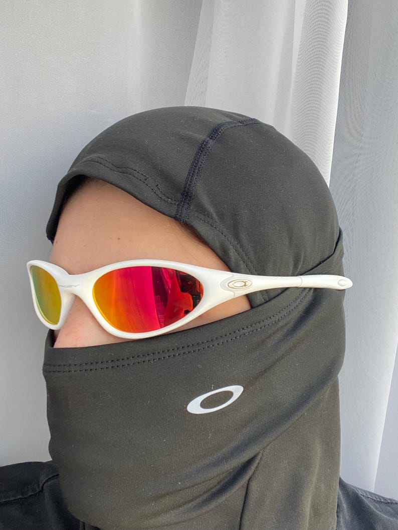 Puede incluir: Gafas de sol con montura blanca y lentes espejadas rojas. Las gafas se llevan con un pasamonta&ntilde;as negro con el logotipo blanco de Oakley. Las lentes reflejan un vibrante tono rojo y amarillo.