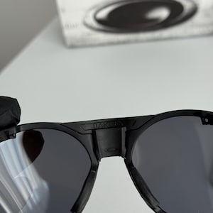 K&ouml;nnte beinhalten: Schwarze Oakley-Sonnenbrille mit runden Gl&auml;sern. Die Sonnenbrille hat einen dicken schwarzen Rahmen, und der Markenname "OAKLEY" ist auf dem oberen B&uuml;gel sichtbar. Die Gl&auml;ser sind dunkelgrau, und das Gesamtdesign ist modern und elegant.