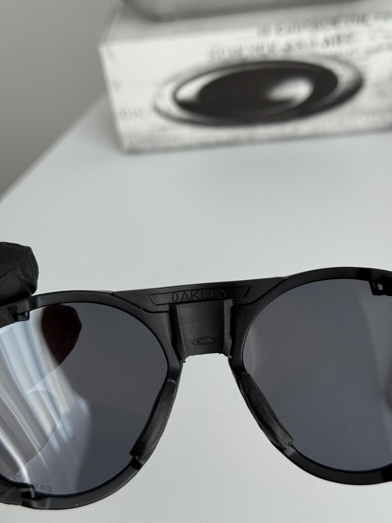 K&ouml;nnte beinhalten: Schwarze Oakley-Sonnenbrille mit runden Gl&auml;sern. Die Sonnenbrille hat einen dicken schwarzen Rahmen, und der Markenname "OAKLEY" ist auf dem oberen B&uuml;gel sichtbar. Die Gl&auml;ser sind dunkelgrau, und das Gesamtdesign ist modern und elegant.