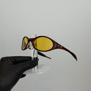 K&ouml;nnte beinhalten: Braune und gelb get&ouml;nte Sonnenbrille mit geschwungenem Rahmendesign. Die Sonnenbrille hat einen dunkelbraunen Rahmen mit gelben Gl&auml;sern. Die B&uuml;gel der Sonnenbrille sind gebogen und erstrecken sich vom Rahmen nach hinten. Die Sonnenbrille wird auf einem transparenten St&auml;nder ausgestellt.