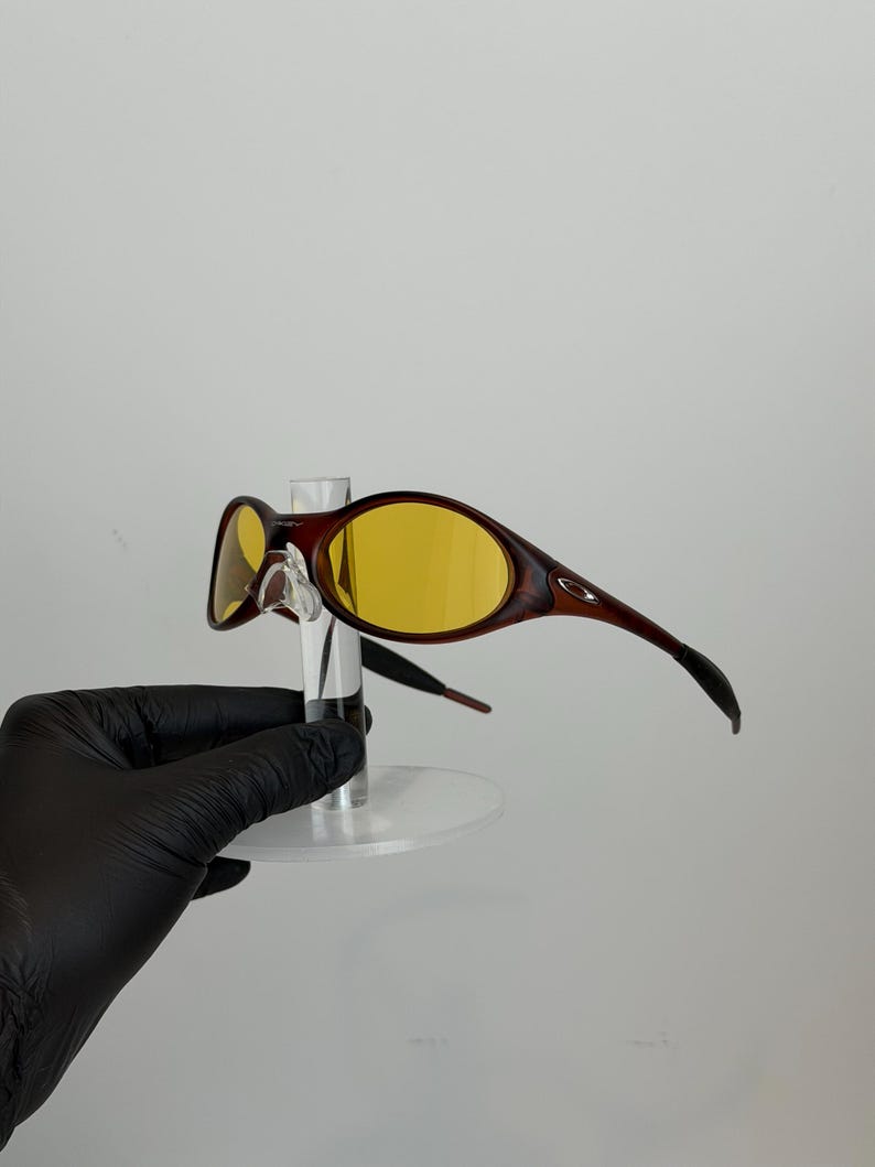 K&ouml;nnte beinhalten: Braune und gelb get&ouml;nte Sonnenbrille mit geschwungenem Rahmendesign. Die Sonnenbrille hat einen dunkelbraunen Rahmen mit gelben Gl&auml;sern. Die B&uuml;gel der Sonnenbrille sind gebogen und erstrecken sich vom Rahmen nach hinten. Die Sonnenbrille wird auf einem transparenten St&auml;nder ausgestellt.