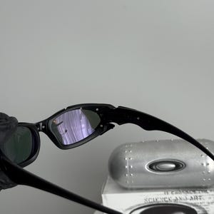 May include: Black sunglasses with a sleek, modern design. The lenses have a reflective, purple hue. The frame is black and angular. The image also shows a silver case with the text "SCIENCE AND ART".