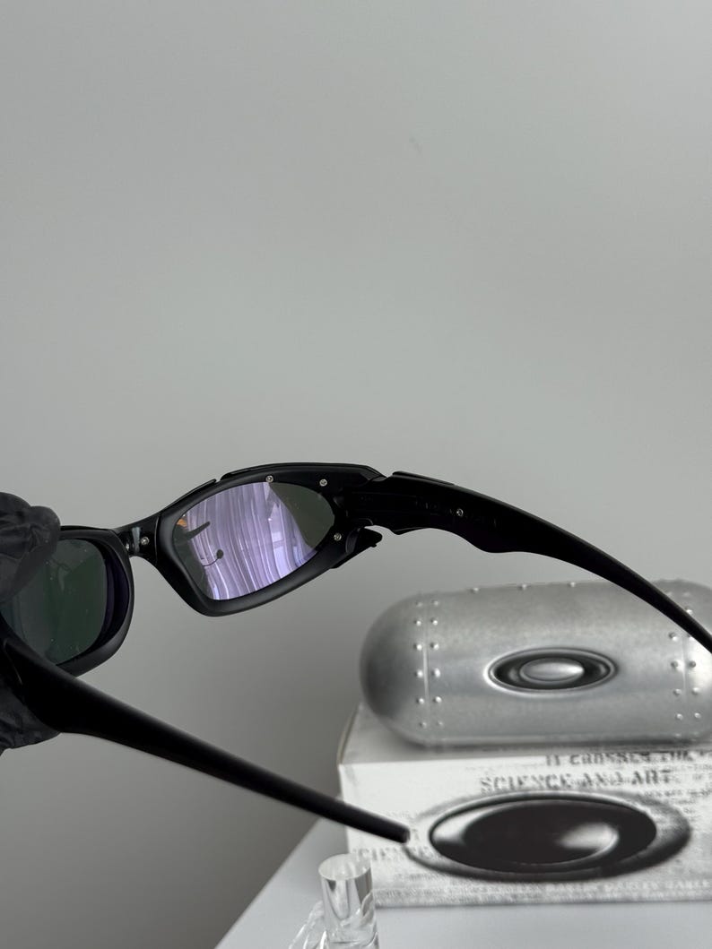 May include: Black sunglasses with a sleek, modern design. The lenses have a reflective, purple hue. The frame is black and angular. The image also shows a silver case with the text "SCIENCE AND ART".