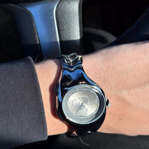 May include: A black Nike watch with a silver face is worn on a wrist. The watch has a unique, sculpted design and a visible Nike logo. The watch face displays the time with multiple dials and hands.