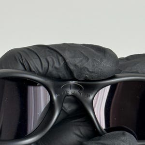 May include: Black sunglasses with a curved frame and dark lenses. The sunglasses have the word "MINUTE" printed on the bridge. The sunglasses are held by a gloved hand.