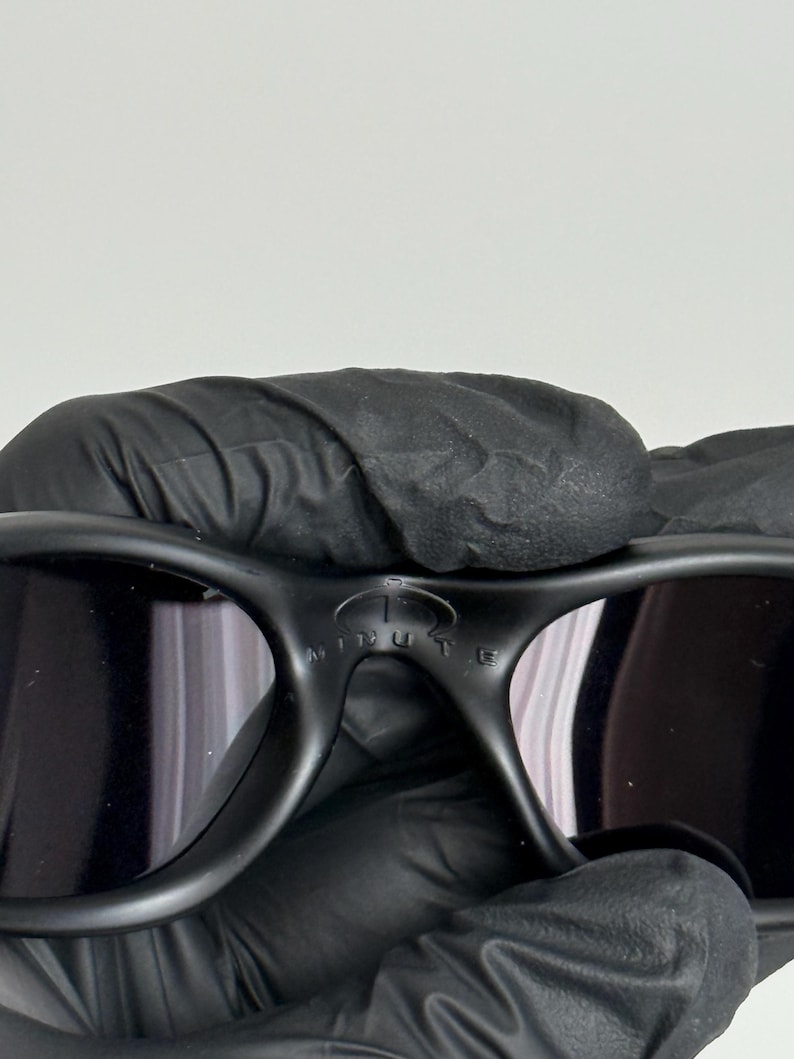 May include: Black sunglasses with a curved frame and dark lenses. The sunglasses have the word "MINUTE" printed on the bridge. The sunglasses are held by a gloved hand.