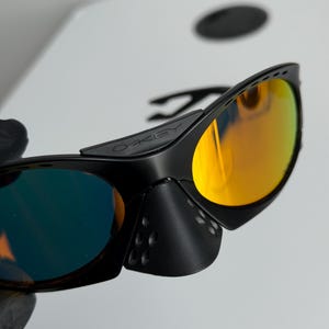 May include: Black sunglasses with orange and blue mirrored lenses. The sunglasses have a sleek, modern design with a black frame and the brand name "OAKLEY" visible on the temple. The lenses reflect a vibrant orange and blue hue.