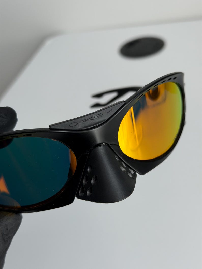 May include: Black sunglasses with orange and blue mirrored lenses. The sunglasses have a sleek, modern design with a black frame and the brand name "OAKLEY" visible on the temple. The lenses reflect a vibrant orange and blue hue.