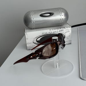 May include: Brown and black sunglasses with a unique, angular design, displayed on a clear stand. A silver case and a box with text are in the background. The sunglasses have brown lenses.
