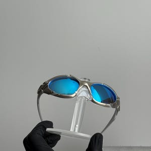 May include: Silver-framed sunglasses with blue lenses. The sunglasses have a unique design with a bridge and temples. The lenses are a vibrant blue color, and the frame has a metallic finish. The sunglasses are displayed on a clear stand.