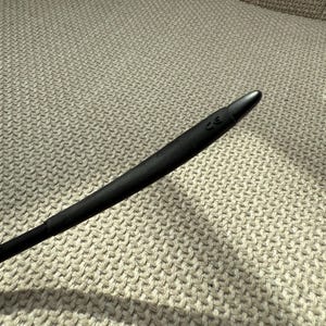 May include: A black, elongated object with a rounded end, possibly a stylus or similar tool. It is set against a textured, light-coloured background, with a subtle shadow. The tip has a small marking.