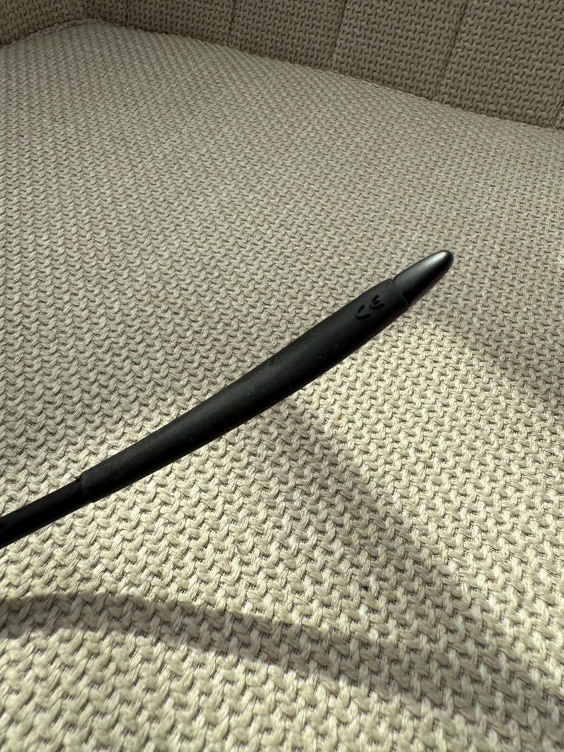 May include: A black, elongated object with a rounded end, possibly a stylus or similar tool. It is set against a textured, light-coloured background, with a subtle shadow. The tip has a small marking.