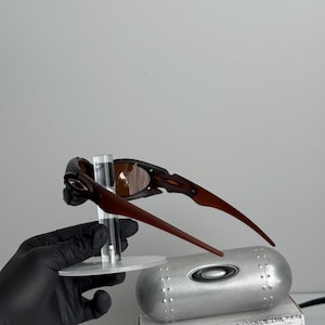 May include: Brown and black sunglasses with a unique frame design. The sunglasses are displayed on a clear stand, with a silver case and a box with the text "SCIENCE AND ART" visible in the background.
