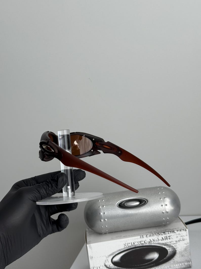 May include: Brown and black sunglasses with a unique frame design. The sunglasses are displayed on a clear stand, with a silver case and a box with the text "SCIENCE AND ART" visible in the background.