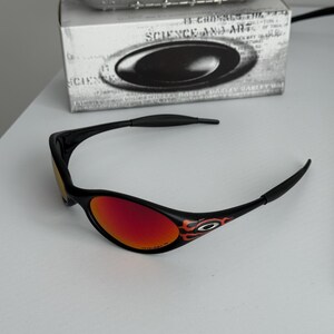 May include: Black sunglasses with red lenses and flame detailing on the temples. The glasses are set against a white background, with a box and a metallic object in the background. The text on the box reads "SCIENCE AND ART".