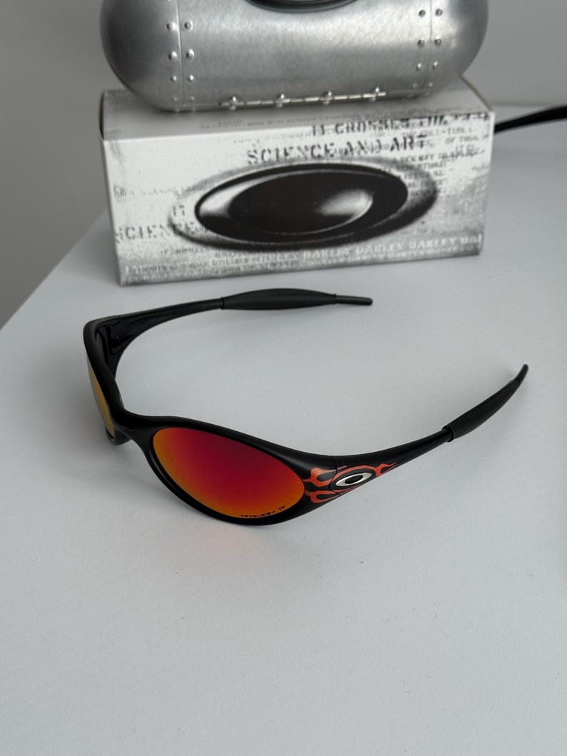 May include: Black sunglasses with red lenses and flame detailing on the temples. The glasses are set against a white background, with a box and a metallic object in the background. The text on the box reads "SCIENCE AND ART".