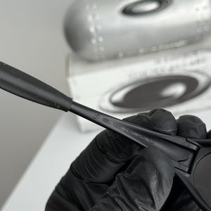 May include: Black sunglasses with the word "jacket" printed on the arm. The sunglasses are held by a gloved hand, with a silver case and a white box in the background.