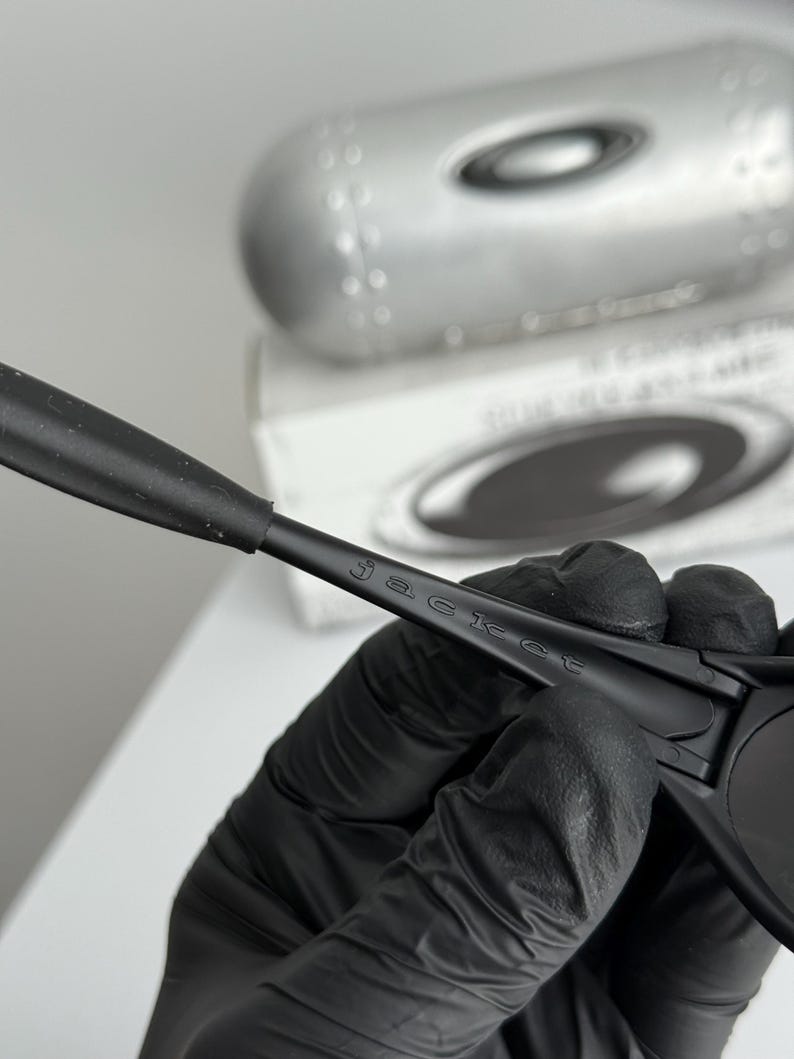 May include: Black sunglasses with the word "jacket" printed on the arm. The sunglasses are held by a gloved hand, with a silver case and a white box in the background.
