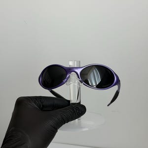 May include: Purple sunglasses with black lenses and a matching frame. The sunglasses are displayed on a clear stand. The arms of the sunglasses are black and curve down. The lenses are reflective.