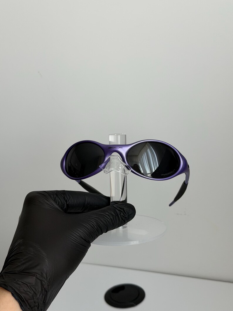 May include: Purple sunglasses with black lenses and a matching frame. The sunglasses are displayed on a clear stand. The arms of the sunglasses are black and curve down. The lenses are reflective.