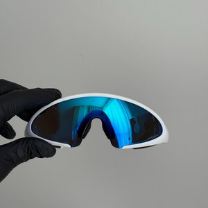 Puede incluir: Gafas de sol deportivas blancas con lentes azules espejadas. Las gafas tienen un dise&ntilde;o curvo y aerodin&aacute;mico y una montura blanca. Las lentes reflejan un color azul brillante, y el estilo general es moderno y deportivo.