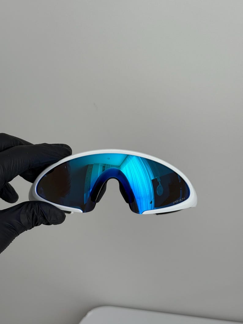 Puede incluir: Gafas de sol deportivas blancas con lentes azules espejadas. Las gafas tienen un dise&ntilde;o curvo y aerodin&aacute;mico y una montura blanca. Las lentes reflejan un color azul brillante, y el estilo general es moderno y deportivo.