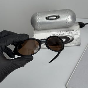 Puede incluir: Gafas de sol Oakley negras con lentes marrones. Las gafas de sol se sostienen frente a un estuche plateado con el logotipo de Oakley. Detr&aacute;s del estuche hay una caja blanca con texto.