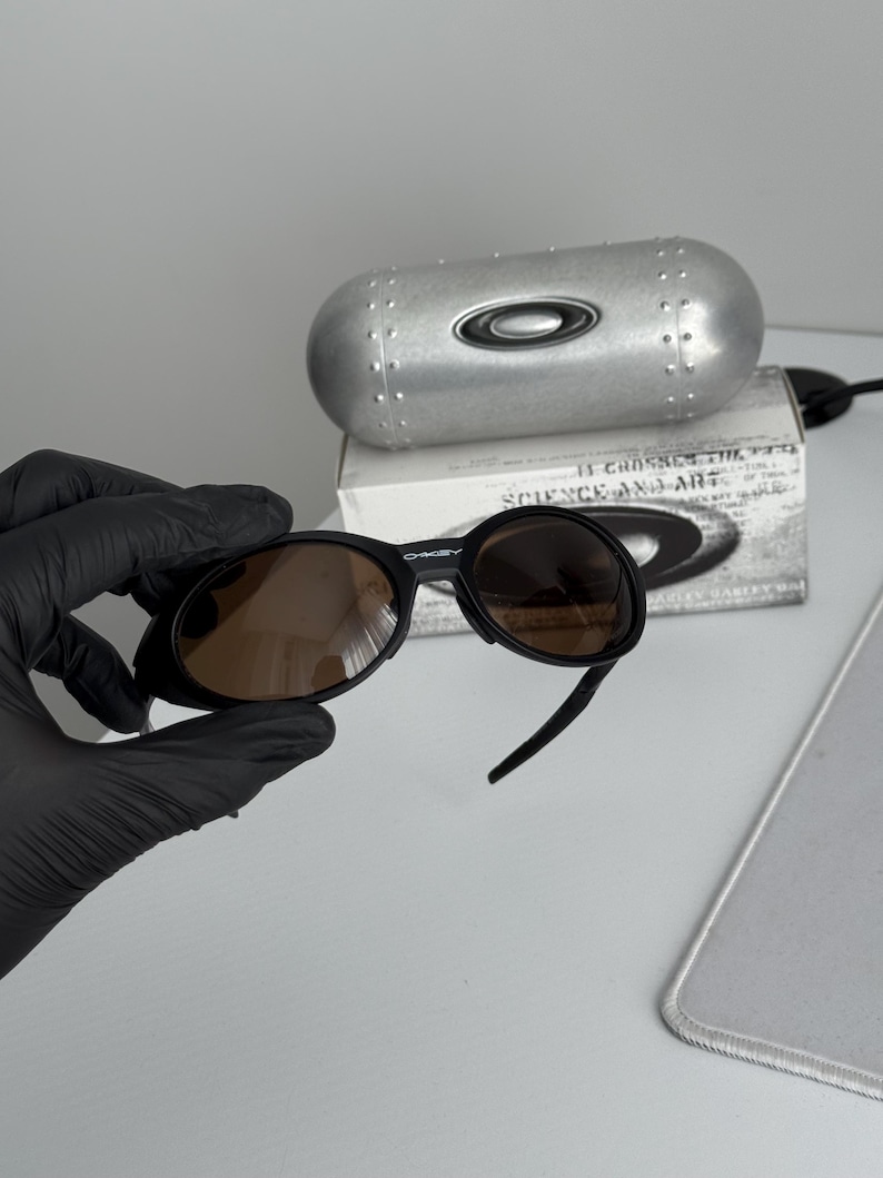 Puede incluir: Gafas de sol Oakley negras con lentes marrones. Las gafas de sol se sostienen frente a un estuche plateado con el logotipo de Oakley. Detr&aacute;s del estuche hay una caja blanca con texto.