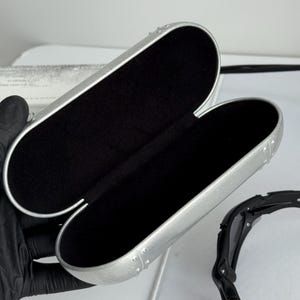 May include: Open silver-colored eyeglasses case with a black interior. The case is oval-shaped and has a riveted design along the edges. Black sunglasses are partially visible to the right of the case.