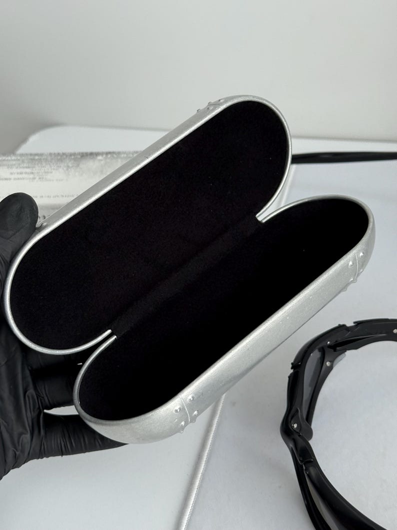 May include: Open silver-colored eyeglasses case with a black interior. The case is oval-shaped and has a riveted design along the edges. Black sunglasses are partially visible to the right of the case.