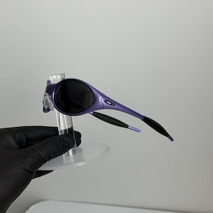 May include: Purple sunglasses with black lenses and black arms. The sunglasses have a sleek, modern design. The frame is a vibrant purple color, and the lenses are dark, providing shade. The arms curve back for a secure fit.