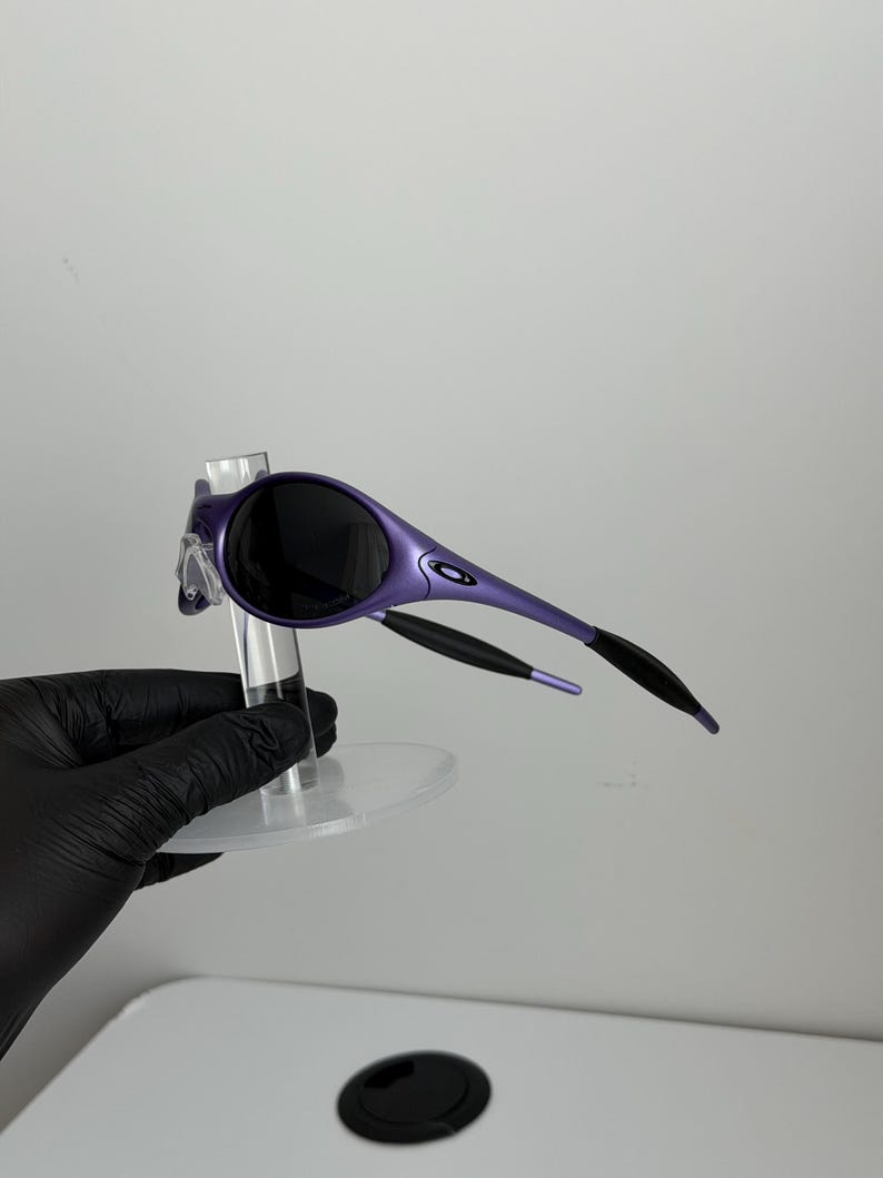 May include: Purple sunglasses with black lenses and black arms. The sunglasses have a sleek, modern design. The frame is a vibrant purple color, and the lenses are dark, providing shade. The arms curve back for a secure fit.