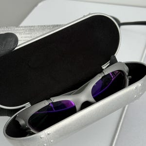 May include: Silver-framed sunglasses with purple lenses rest inside a matching silver case with a black interior. The case is open, revealing the sunglasses. The sunglasses have a sleek, modern design.