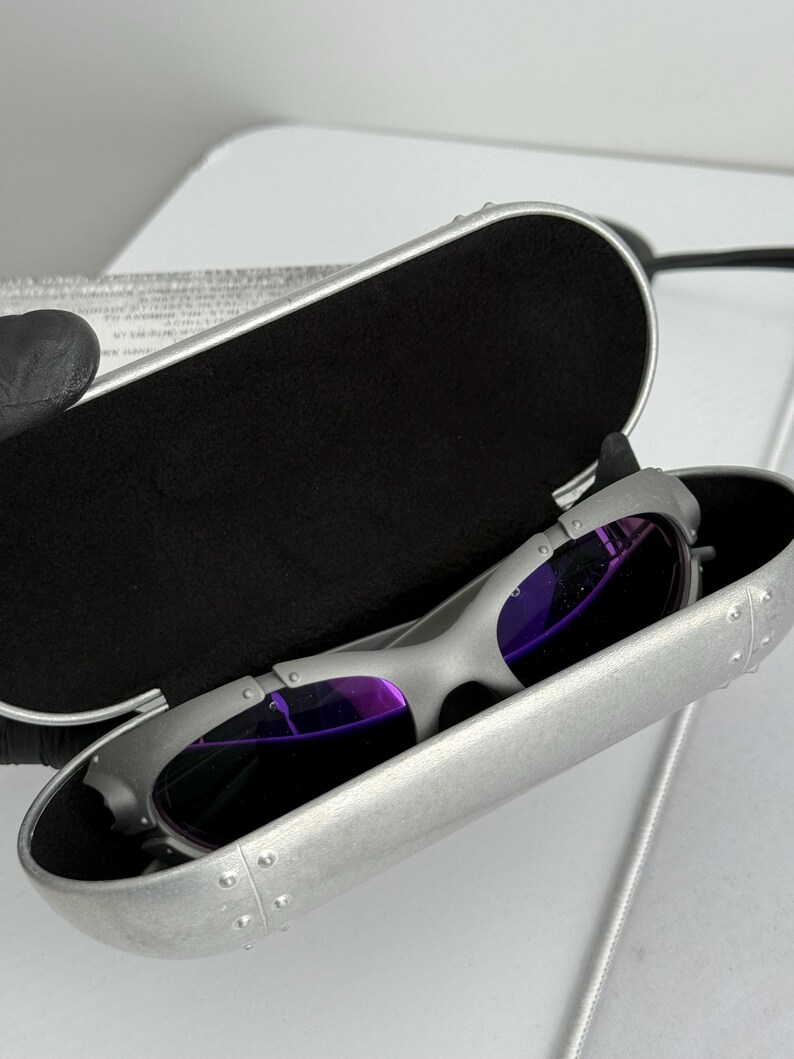 May include: Silver-framed sunglasses with purple lenses rest inside a matching silver case with a black interior. The case is open, revealing the sunglasses. The sunglasses have a sleek, modern design.