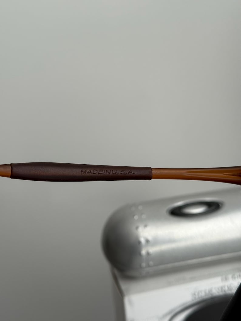 May include: Close-up of brown eyeglasses with the text "MADE IN U.S.A." on the temple. The frame is a light brown color, and the temple has a darker brown grip.