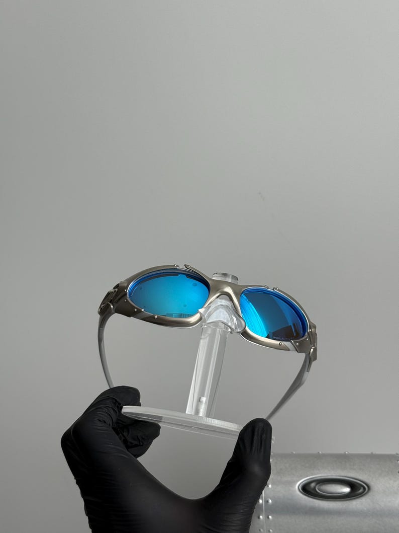May include: Silver-framed sunglasses with blue lenses. The sunglasses have a unique design with a bridge and temples. The lenses are a vibrant blue color, and the frame has a metallic finish. The sunglasses are displayed on a clear stand.