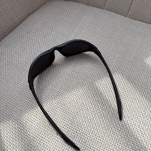 May include: Black sunglasses with a streamlined design. The sunglasses feature a curved frame and dark lenses. The arms are thin and extend back, creating a modern look. The sunglasses are resting on a textured, light-coloured surface.