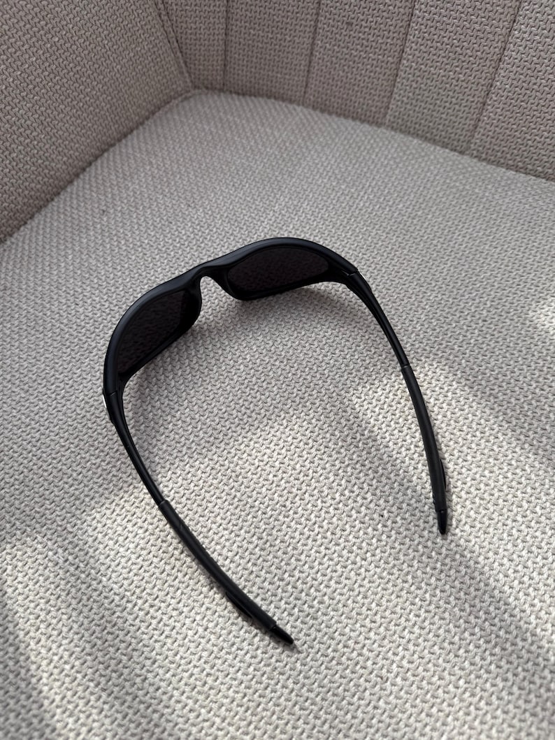 May include: Black sunglasses with a streamlined design. The sunglasses feature a curved frame and dark lenses. The arms are thin and extend back, creating a modern look. The sunglasses are resting on a textured, light-coloured surface.