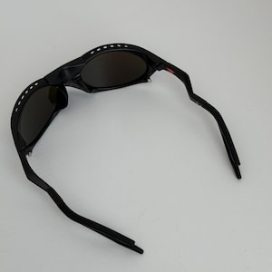May include: Black sunglasses with a curved frame and dark lenses. The sunglasses have a series of white dots along the top of the frame and a red accent on the right arm. The arms are curved and angled.